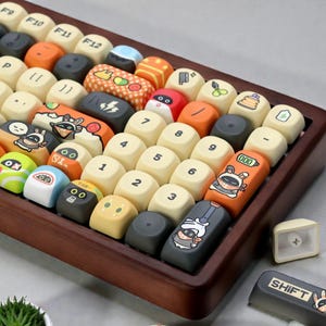 May include: A custom keyboard with various keycap designs. The keycaps feature a mix of colors, including orange, blue, and yellow, with cartoon illustrations and numbers. The keyboard is set in a wooden frame.