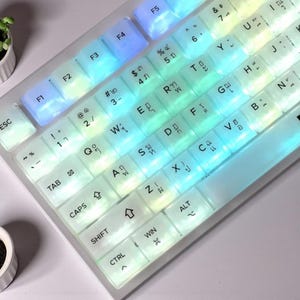 May include: A backlit keyboard with transparent keycaps. The keys are illuminated with a gradient of colors, transitioning from blue to green and yellow. The keyboard has a standard layout with function keys and special characters.