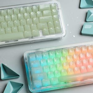May include: Two mechanical keyboards with transparent cases. One keyboard has light green keycaps, the other has rainbow-colored keycaps. The keyboards are on a light gray surface, with teal-colored geometric shapes scattered around.