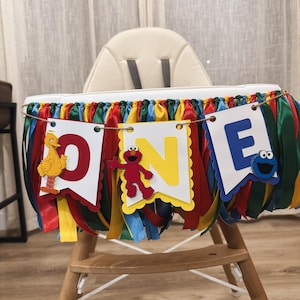 May include: A high chair decorated with a colorful banner that reads "ONE" with Sesame Street characters. The banner has red, yellow, blue, and green ribbons. The letters are on white and yellow cutouts.