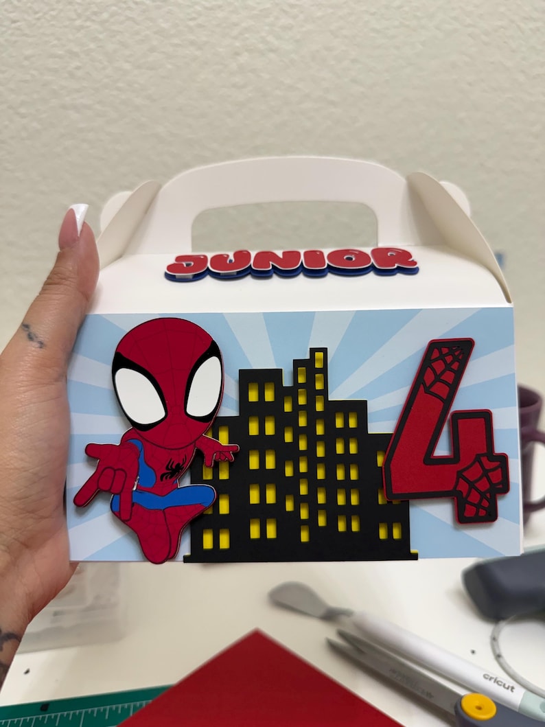 Op de afbeelding: Een witte zadeldakdoos met een handvat, met een Spider-Man-ontwerp. De doos heeft een rood en blauw Spider-Man figuur, een zwart stadsbeeld en het rode nummer 4. Het woord "JUNIOR" staat bovenaan.