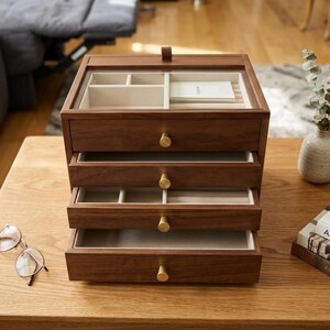May include: A wooden jewelry box with four drawers and a clear glass top. The box is made of dark brown wood and has gold-colored knobs. The drawers are open, revealing beige compartments. A pair of glasses and a book are on the table.