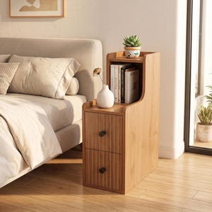 May include: A light brown wooden bedside table featuring two drawers with vertical fluted detailing and black circular knobs. The unit includes an open shelf for books and a top surface holding a small potted succulent and a white ceramic vase.