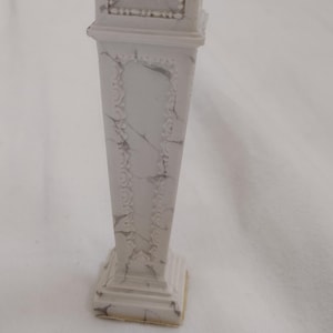 Puede incluir: Una columna decorativa blanca con un patrón similar al mármol. La columna tiene una base cuadrada, un cuerpo rectangular y una pequeña parte superior. La columna mide aproximadamente 15 cm de altura.