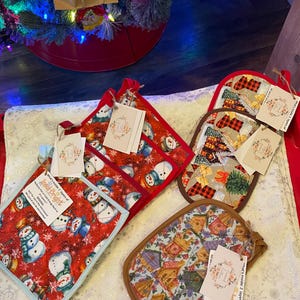 Könnte beinhalten: Eine Sammlung von Topflappen mit Weihnachtsmotiven, darunter Schneemänner, Patchwork und Feiertagsmuster. Jeder Topflappen hat ein angebrachtes Etikett. Der Text "A savior has been born to you" ist im Hintergrund sichtbar.