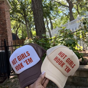 May include: Two trucker hats featuring the text "HOT GIRLS HOOK 'EM" in burnt orange lettering. One hat has a white front panel with a dark brown brim and mesh back, while the other is a solid light beige color with a matching mesh back panel.