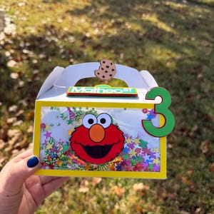 May include: A white party favor box with a yellow frame, featuring Elmo's face and the number 3. The box is decorated with colorful confetti and a cookie. The name "Matheos" is written on the box.