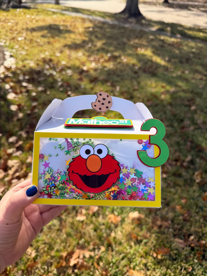 May include: A white party favor box with a yellow frame, featuring Elmo's face and the number 3. The box is decorated with colorful confetti and a cookie. The name "Matheos" is written on the box.