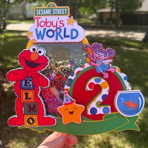 May include: A colorful Sesame Street-themed birthday cake topper. Features Elmo, Abby Cadabby, a star, and a fishbowl. The topper includes the text "Toby's World" and the number "2".