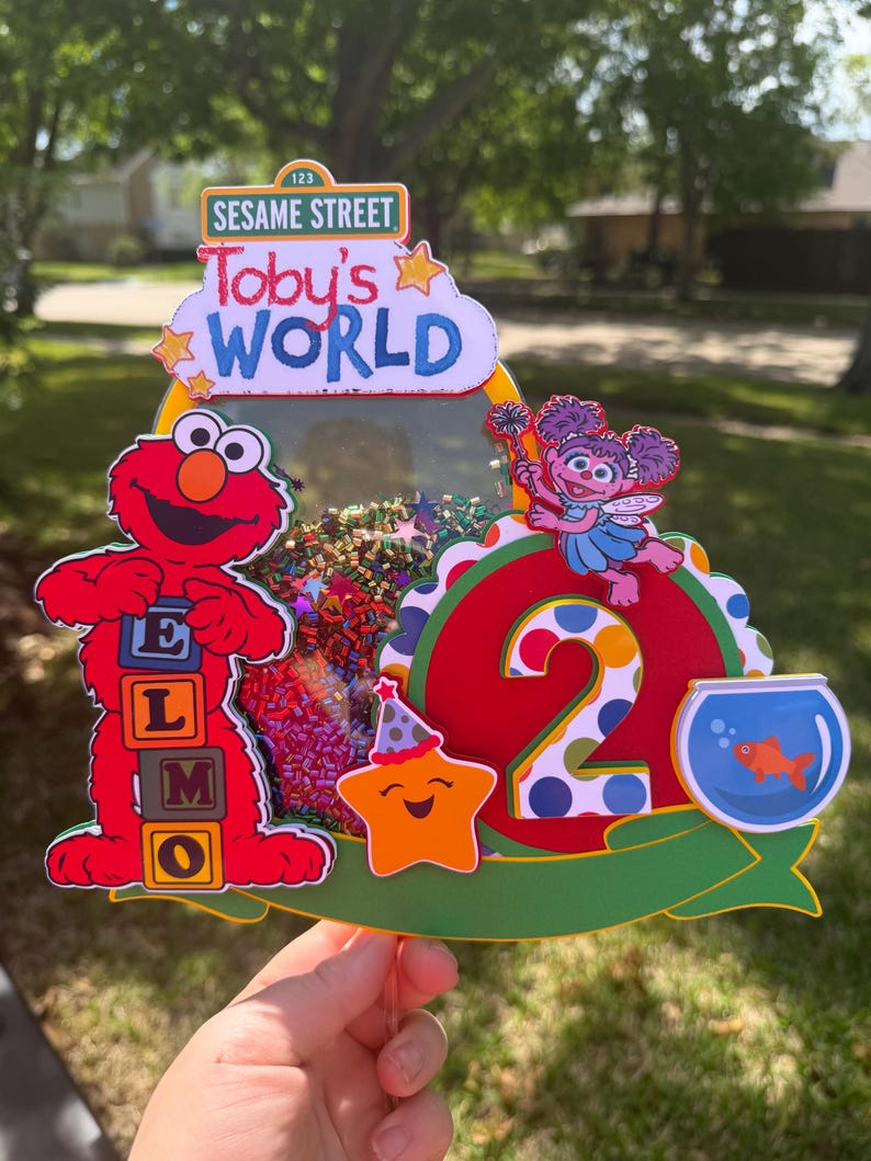 May include: A colorful Sesame Street-themed birthday cake topper. Features Elmo, Abby Cadabby, a star, and a fishbowl. The topper includes the text "Toby's World" and the number "2".