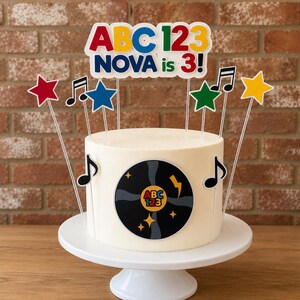 May include: A white frosted cake topped with a colorful sign reading ABC 123 NOVA is 3! and various cake toppers including red, blue, green, and yellow stars, black musical notes, and a central black vinyl record graphic with ABC 123 text.