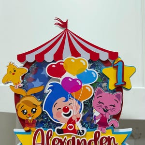Puede incluir: Un adorno para tarta colorido con tem&aacute;tica de circo con el nombre "Alexander". Presenta personajes de dibujos animados, globos y el n&uacute;mero uno. El adorno tiene un dise&ntilde;o de carpa a rayas rojas y blancas, ideal para un cumplea&ntilde;os.