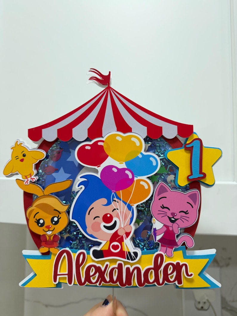 Puede incluir: Un adorno para tarta colorido con tem&aacute;tica de circo con el nombre "Alexander". Presenta personajes de dibujos animados, globos y el n&uacute;mero uno. El adorno tiene un dise&ntilde;o de carpa a rayas rojas y blancas, ideal para un cumplea&ntilde;os.