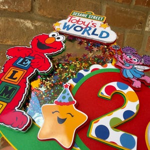 May include: A colorful Sesame Street-themed birthday decoration featuring Elmo, Abby Cadabby, and a smiling star. The design includes the words "Toby's World" and the number "2". The decoration is primarily red, yellow, green, and blue.