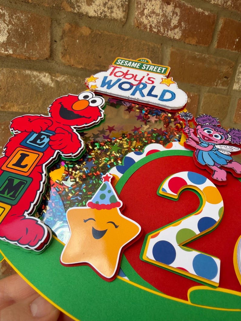 May include: A colorful Sesame Street-themed birthday decoration featuring Elmo, Abby Cadabby, and a smiling star. The design includes the words "Toby's World" and the number "2". The decoration is primarily red, yellow, green, and blue.