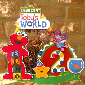 May include: A colorful Sesame Street-themed birthday decoration featuring Elmo, Abby Cadabby, and a smiling star. The design includes the text "Toby's WORLD" and the number "2". The decoration is filled with confetti and stars.