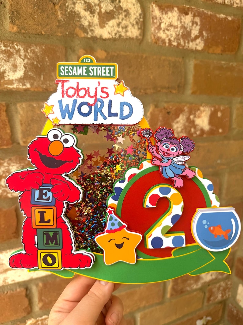 May include: A colorful Sesame Street-themed birthday decoration featuring Elmo, Abby Cadabby, and a smiling star. The design includes the text "Toby's WORLD" and the number "2". The decoration is filled with confetti and stars.
