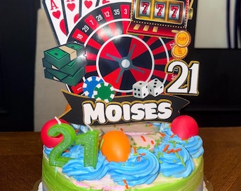 May include: A round cake with blue and green frosting, topped with a casino-themed topper featuring playing cards, a roulette wheel, a slot machine with 777, dice, poker chips, and a stack of cash. A black banner displays the name MOISES and the number 21.