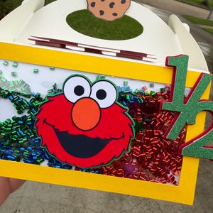 May include: A white and yellow box with a cutout and a cookie cutout on top. The box features a red cartoon character face, colorful confetti, and the number "1/2" in green glitter. The box is likely for a birthday or celebration.
