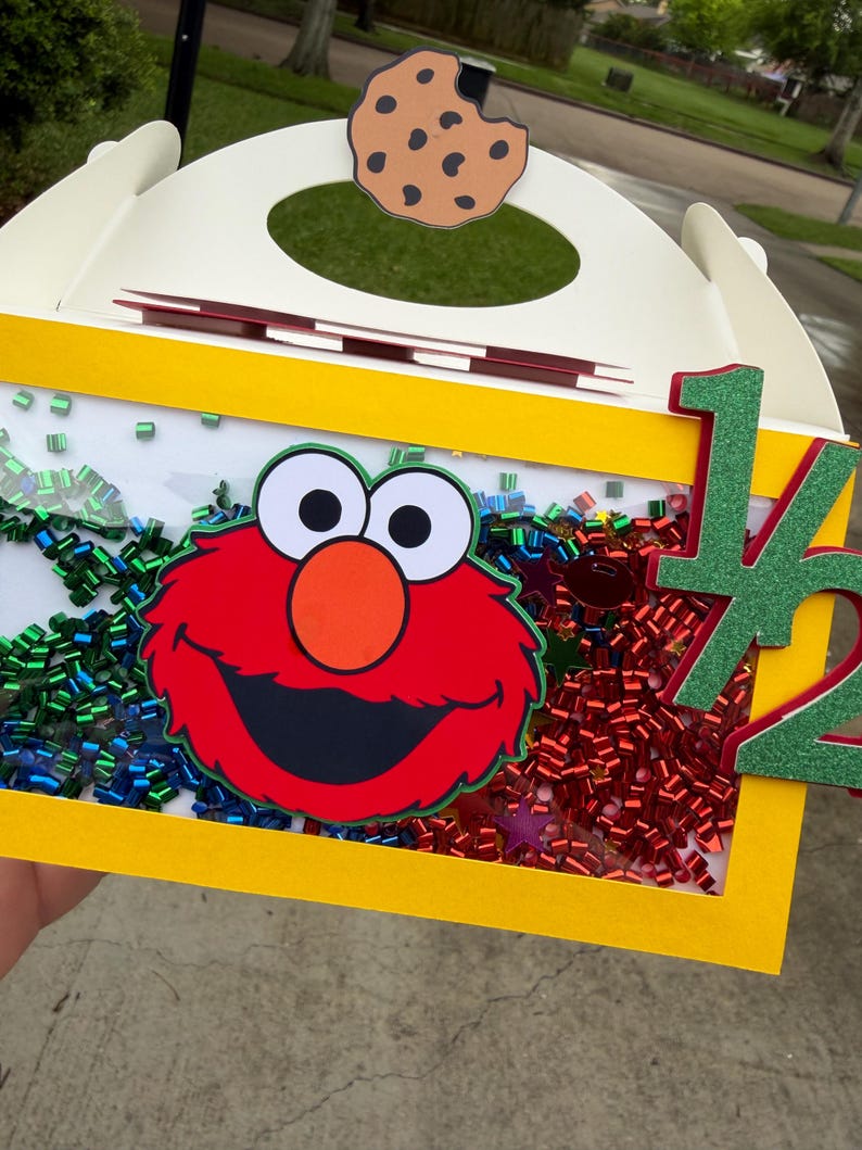 May include: A white and yellow box with a cutout and a cookie cutout on top. The box features a red cartoon character face, colorful confetti, and the number "1/2" in green glitter. The box is likely for a birthday or celebration.