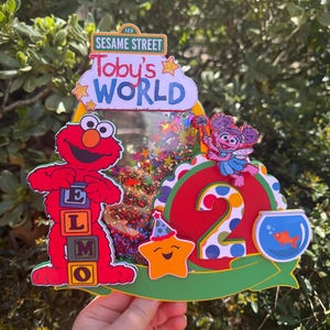 May include: A colorful Sesame Street-themed cake topper featuring Elmo, Abby Cadabby, and a smiling star. The topper includes the text "Toby's WORLD" and the number "2", with a fishbowl and confetti accents. The background is a blurred green.