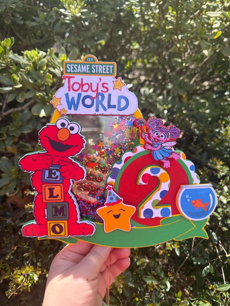 May include: A colorful Sesame Street-themed cake topper featuring Elmo, Abby Cadabby, and a smiling star. The topper includes the text "Toby's WORLD" and the number "2", with a fishbowl and confetti accents. The background is a blurred green.
