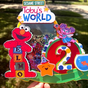 May include: A colorful Sesame Street-themed birthday cake topper. It features Elmo holding letter blocks, the number 2, and the text "Toby's WORLD". The topper includes a star, a fishbowl, and confetti.