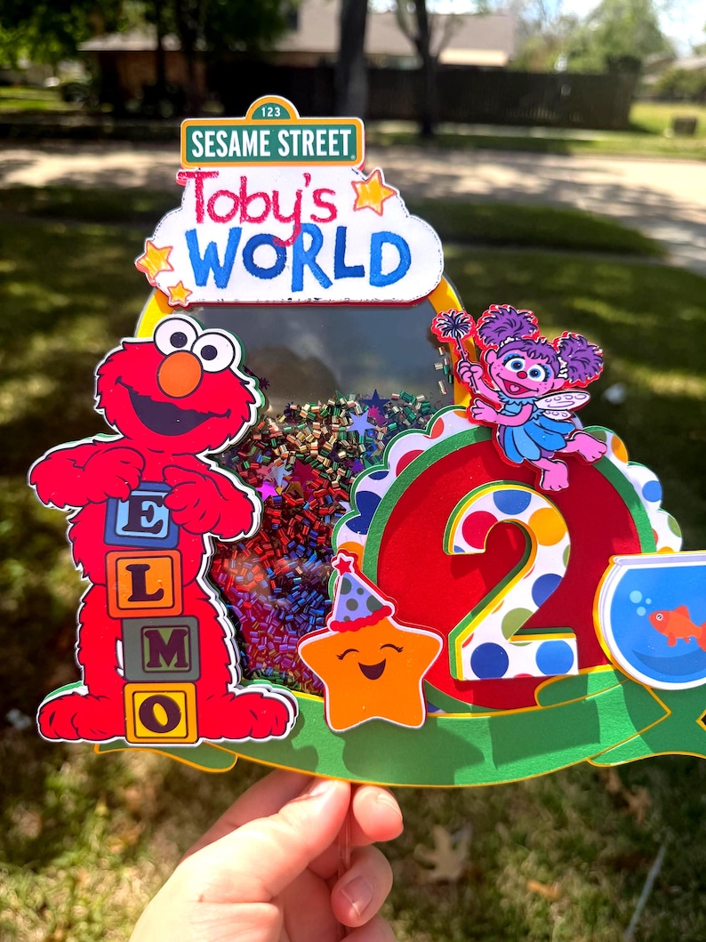May include: A colorful Sesame Street-themed birthday cake topper. It features Elmo holding letter blocks, the number 2, and the text "Toby's WORLD". The topper includes a star, a fishbowl, and confetti.