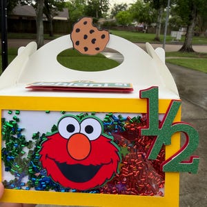 May include: A yellow box with a clear front panel featuring a red Elmo character and colorful confetti. A cookie cutout is on top, and a green and red "1/2" is on the side. The box is decorated with a Sesame Street theme.