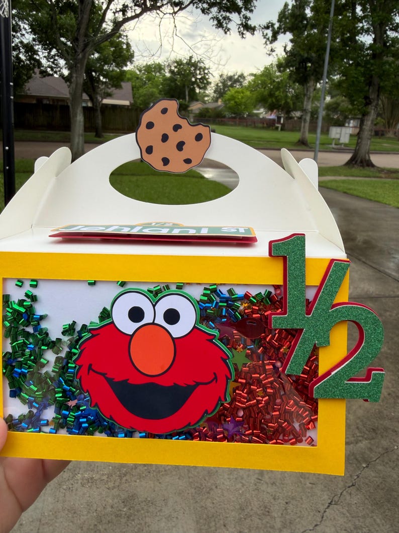 May include: A yellow box with a clear front panel featuring a red Elmo character and colorful confetti. A cookie cutout is on top, and a green and red "1/2" is on the side. The box is decorated with a Sesame Street theme.