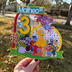 May include: A colorful birthday cake topper featuring Sesame Street characters, the number 3, and the text "Matheo ST." The topper includes Big Bird, Elmo, Cookie Monster, and other beloved characters, with a green banner and a cookie.