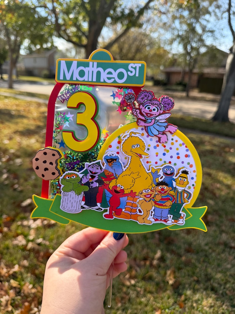 May include: A colorful birthday cake topper featuring Sesame Street characters, the number 3, and the text "Matheo ST." The topper includes Big Bird, Elmo, Cookie Monster, and other beloved characters, with a green banner and a cookie.