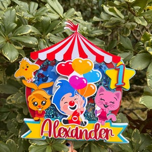 Puede incluir: Un adorno para pastel colorido con tem&aacute;tica de circo con el nombre "Alexander" en escritura roja. El adorno presenta personajes de dibujos animados, globos y el n&uacute;mero "1" en forma de estrella, todo sobre una carpa de circo a rayas rojas y blancas.