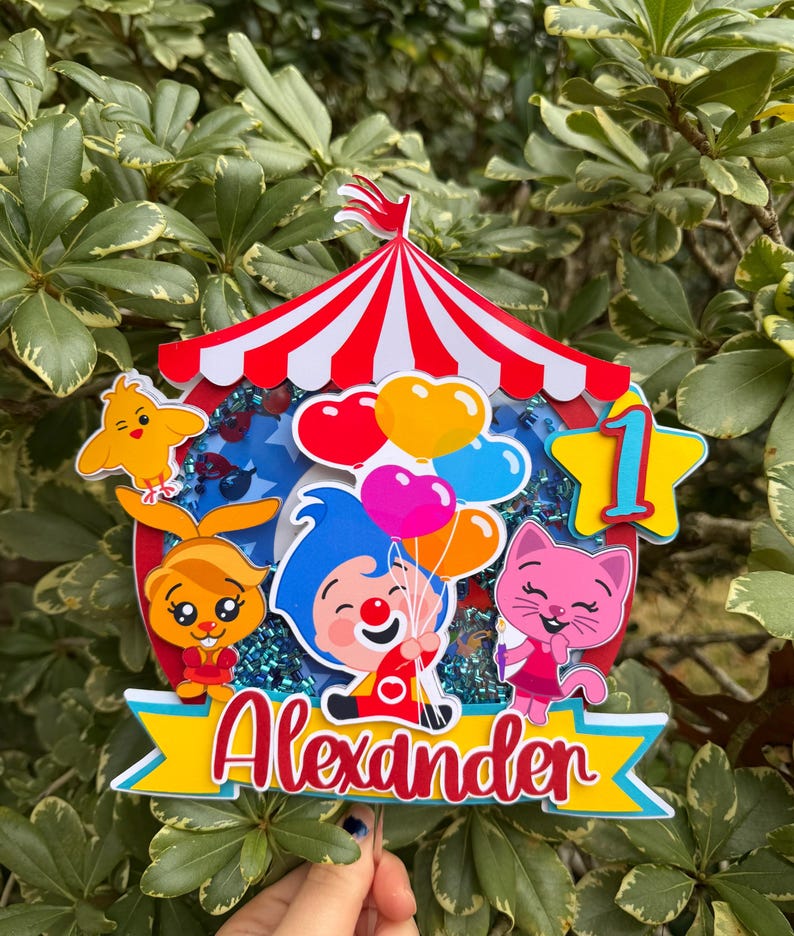 Puede incluir: Un adorno para pastel colorido con tem&aacute;tica de circo con el nombre "Alexander" en escritura roja. El adorno presenta personajes de dibujos animados, globos y el n&uacute;mero "1" en forma de estrella, todo sobre una carpa de circo a rayas rojas y blancas.