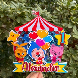 Puede incluir: Un adorno para pastel con tem&aacute;tica de circo colorido con un dise&ntilde;o de carpa a rayas rojas y blancas. Presenta personajes de dibujos animados, globos y el nombre "Alexander" en escritura roja. Tambi&eacute;n se incluye una estrella amarilla con el n&uacute;mero "1".