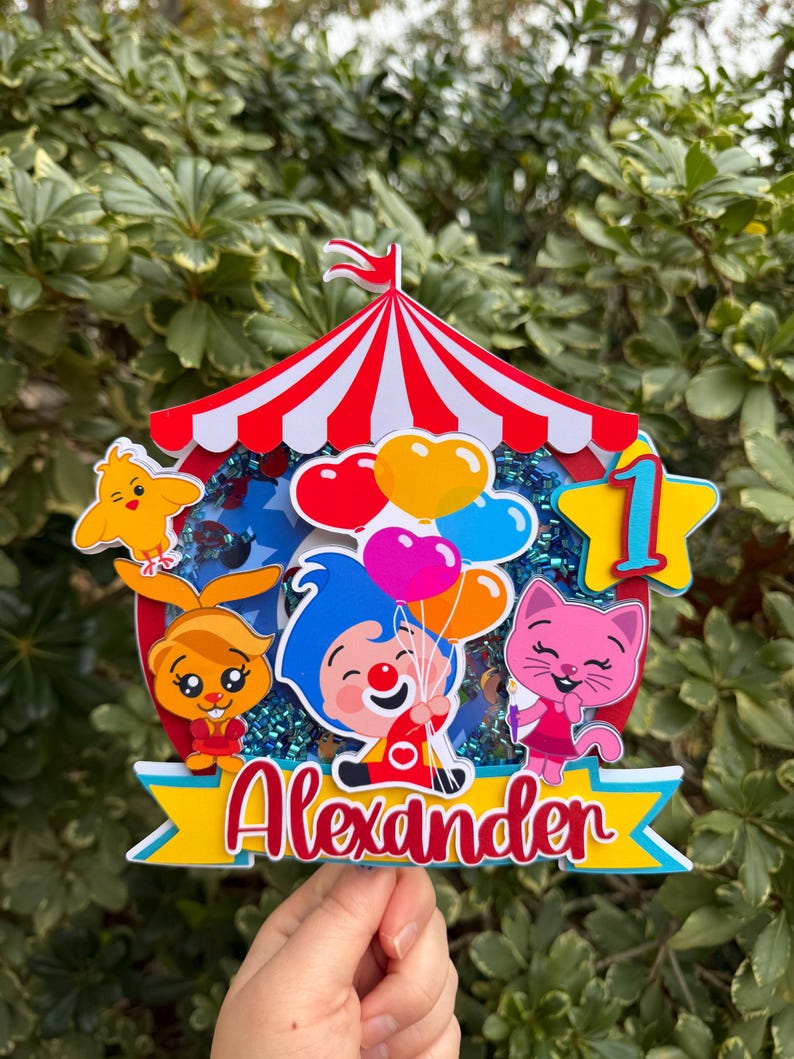 Puede incluir: Un adorno para pastel con tem&aacute;tica de circo colorido con un dise&ntilde;o de carpa a rayas rojas y blancas. Presenta personajes de dibujos animados, globos y el nombre "Alexander" en escritura roja. Tambi&eacute;n se incluye una estrella amarilla con el n&uacute;mero "1".