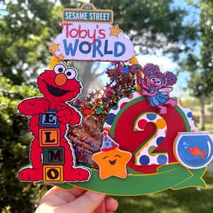 May include: A colorful Sesame Street-themed birthday cake topper. Features Elmo with letter blocks, the number 2, a star, and a fishbowl. The topper includes the text "Toby's World" and "Sesame Street".