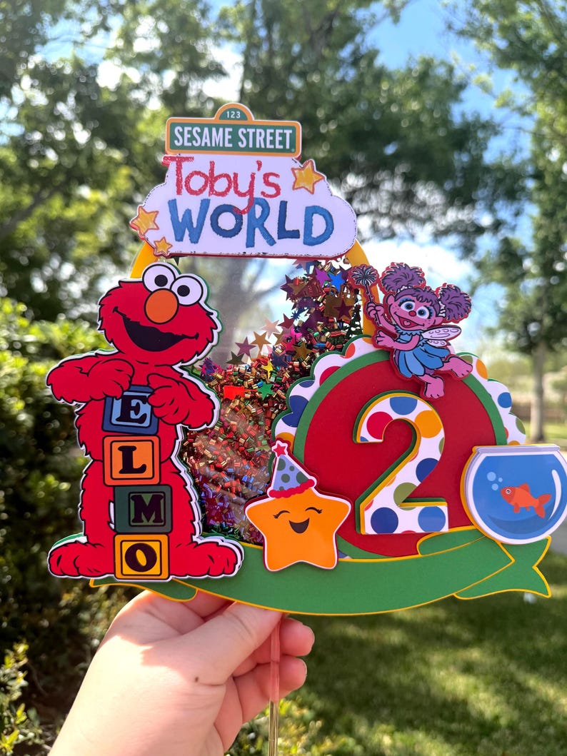 May include: A colorful Sesame Street-themed birthday cake topper. Features Elmo with letter blocks, the number 2, a star, and a fishbowl. The topper includes the text "Toby's World" and "Sesame Street".