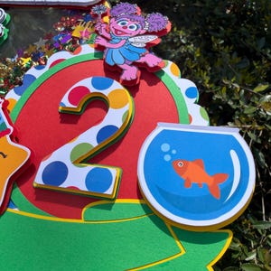 May include: A colorful Sesame Street-themed birthday decoration featuring the text "Toby's WORLD". The design includes a large number "2" with polka dots, a cartoon character, and a goldfish in a bowl. The color scheme includes red, green, blue, and yellow.
