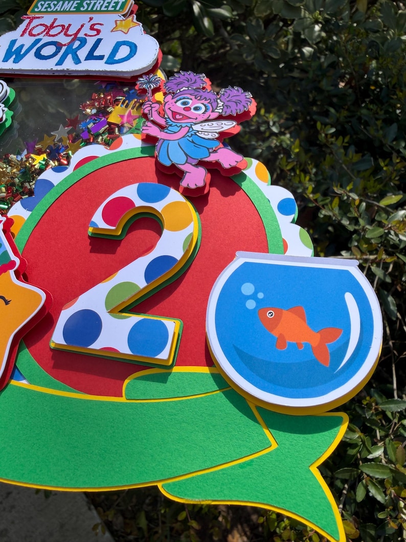 May include: A colorful Sesame Street-themed birthday decoration featuring the text "Toby's WORLD". The design includes a large number "2" with polka dots, a cartoon character, and a goldfish in a bowl. The color scheme includes red, green, blue, and yellow.