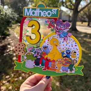 May include: A colorful cake topper featuring Sesame Street characters, the number 3, and the text "Matheo ST". The design includes Big Bird, Elmo, Cookie Monster, and other beloved characters, perfect for a birthday celebration.