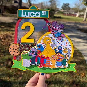 May include: A colorful birthday cake topper featuring Sesame Street characters. The topper includes a large yellow number 2, the street sign "Luca ST", and images of Cookie Monster, Big Bird, Elmo, and other characters. The background is a mix of confetti.