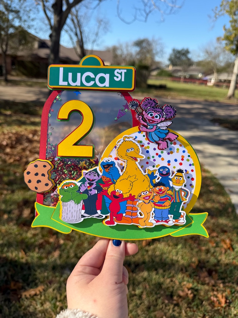 May include: A colorful birthday cake topper featuring Sesame Street characters. The topper includes a large yellow number 2, the street sign "Luca ST", and images of Cookie Monster, Big Bird, Elmo, and other characters. The background is a mix of confetti.
