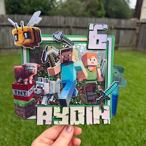 May include: A Minecraft-themed cake topper featuring a square frame with green borders, filled with green glitter. It displays characters Steve and Alex, a bee, a pink sheep, a wolf, a TNT block, a grass block, and the name AYDIN in block letters.