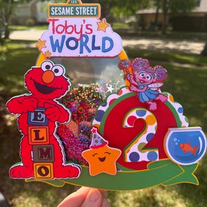 May include: A colorful Sesame Street-themed birthday cake topper. It features Elmo, Abby Cadabby, a smiling star, and a goldfish in a bowl. The topper includes the text "Toby's World" and the number "2".