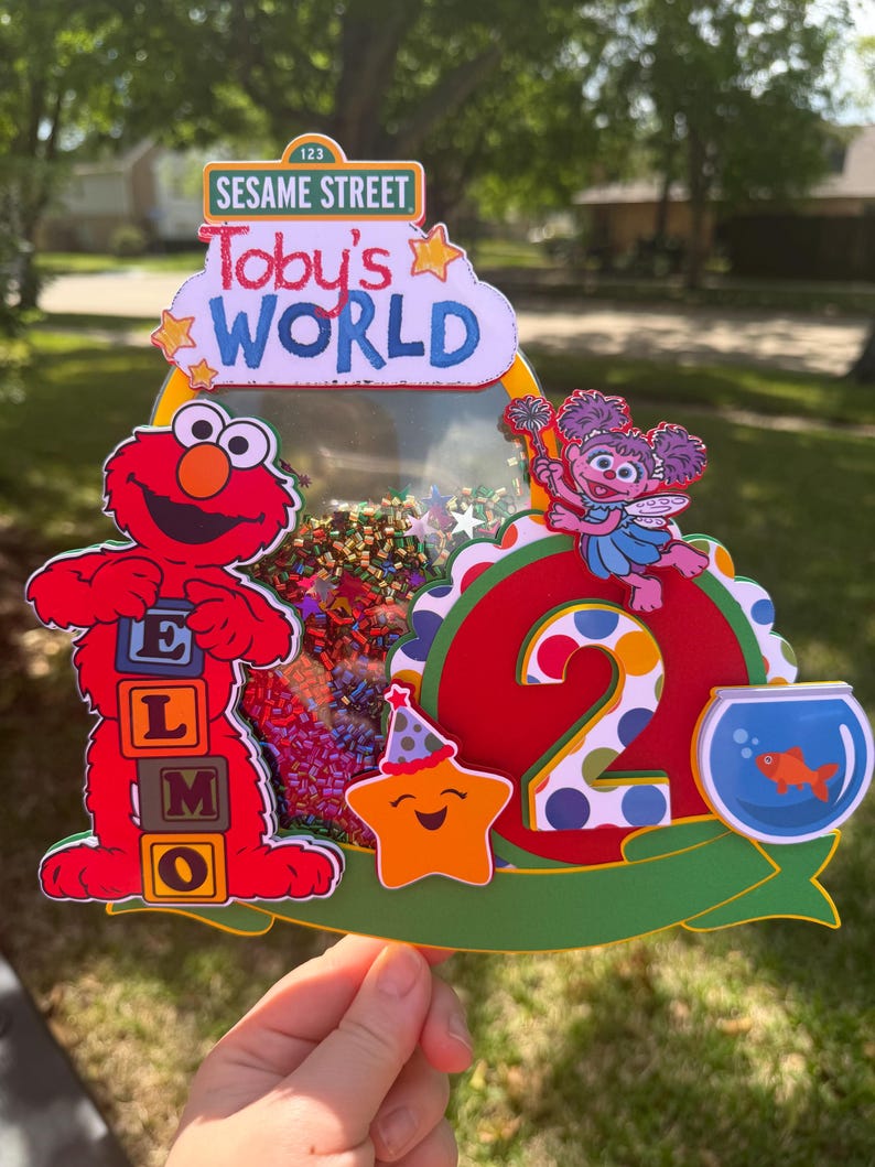 May include: A colorful Sesame Street-themed birthday cake topper. It features Elmo, Abby Cadabby, a smiling star, and a goldfish in a bowl. The topper includes the text "Toby's World" and the number "2".