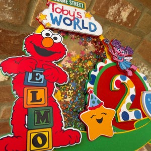 May include: A colorful Sesame Street-themed birthday decoration featuring Elmo holding letter blocks spelling "ELMO". The decoration includes the text "Toby's World" and a smiling star with a party hat. The number "2" is also visible.