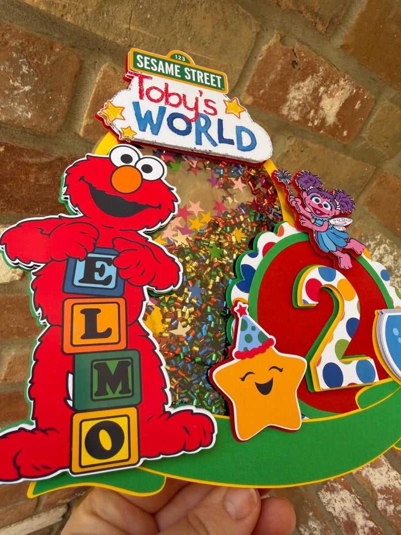 May include: A colorful Sesame Street-themed birthday decoration featuring Elmo holding letter blocks spelling "ELMO". The decoration includes the text "Toby's World" and a smiling star with a party hat. The number "2" is also visible.