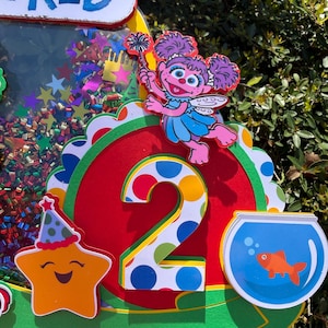 May include: A colorful birthday decoration featuring a number 2, a smiling star with a party hat, and a cartoon character holding a wand. The decoration includes a fishbowl with a goldfish and the text "Baby's World".