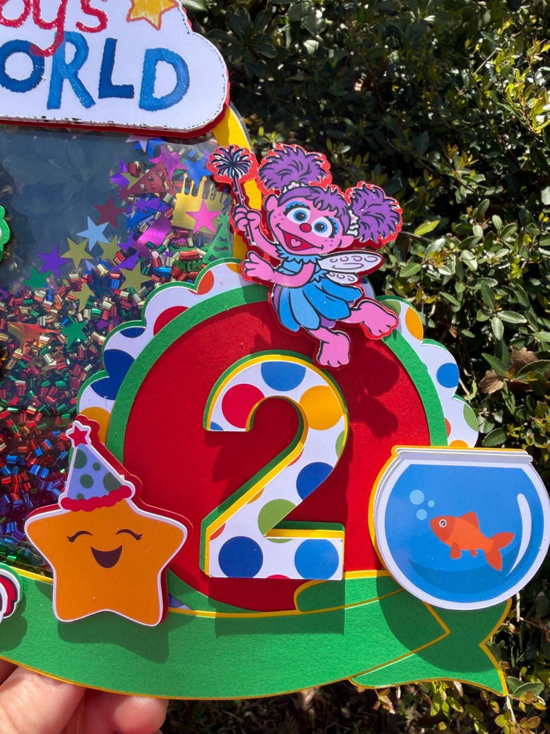 May include: A colorful birthday decoration featuring a number 2, a smiling star with a party hat, and a cartoon character holding a wand. The decoration includes a fishbowl with a goldfish and the text "Baby's World".